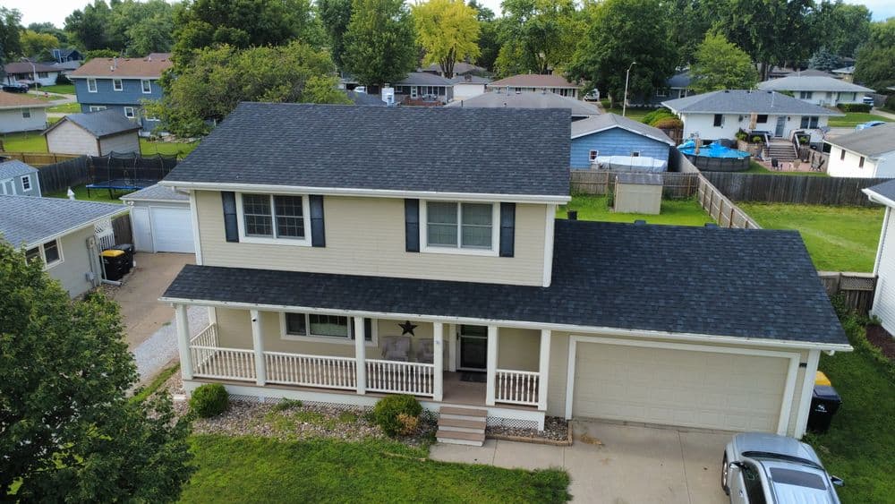 Two-story beige house with a dark roof, green lawn, and driveway in a suburban neighborhood.