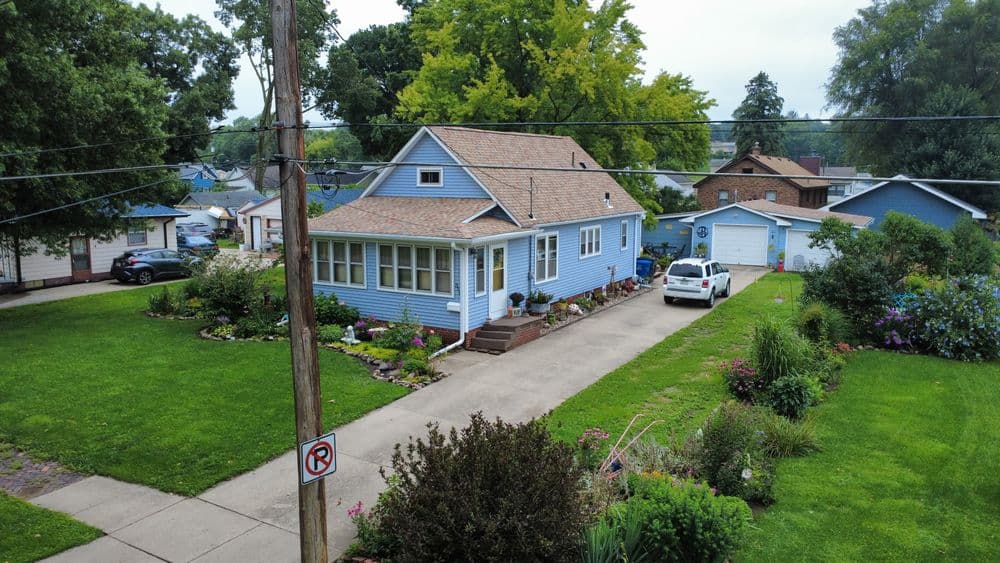 Blue house with garden and driveway, surrounded by green lawn and trees in a residential area.