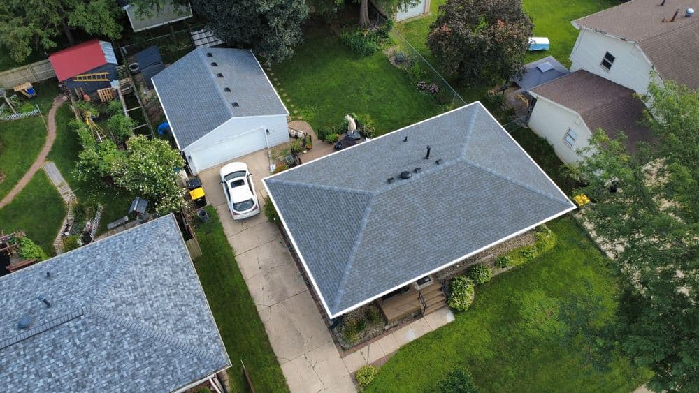 Aerial view of residential rooftops with green lawns and parked cars on a sunny day.
