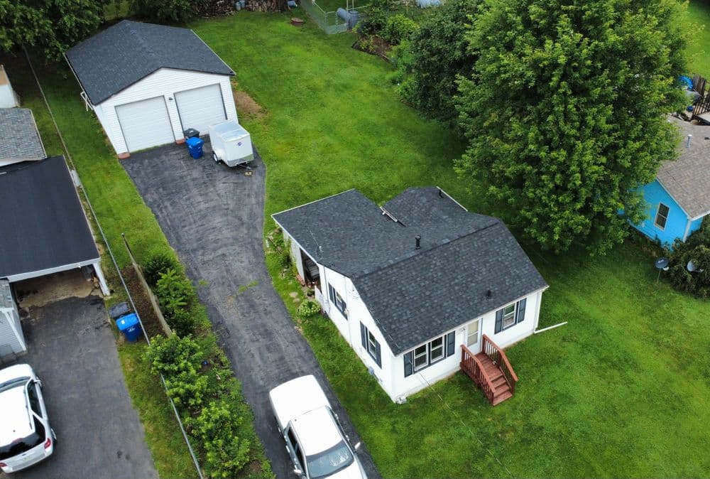 Aerial view of a small white house with a black roof, surrounded by green lawn and garages.