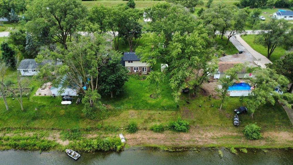 Aerial view of a house near a river, surrounded by trees and outdoor recreational spaces.