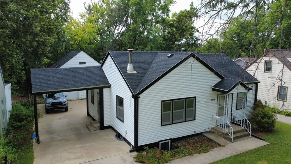 Aerial view of a charming white house with a black roof and carport in a green neighborhood.