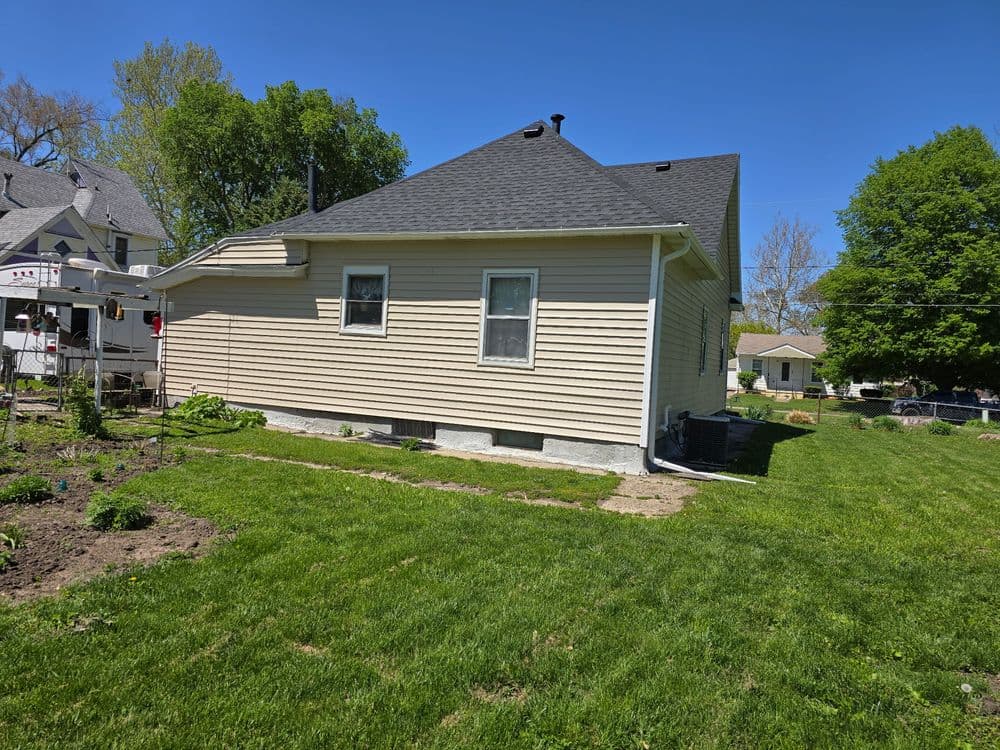 Side view of a beige house with a shingled roof, surrounded by a green lawn and garden.