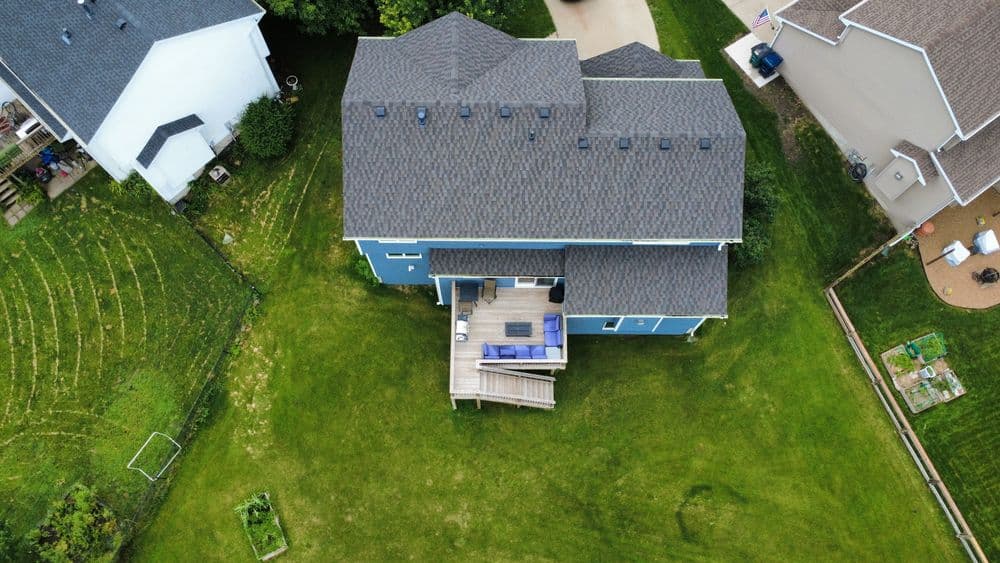 Aerial view of a blue house with a wooden deck surrounded by lush green grass and gardens.
