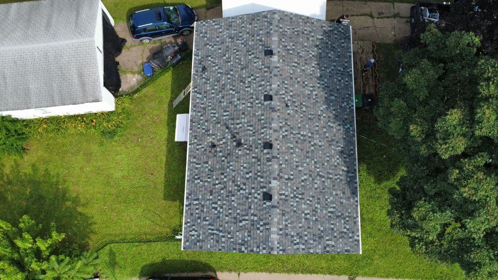 Aerial view of a residential roof with shingles, surrounded by green lawn and nearby vehicles.