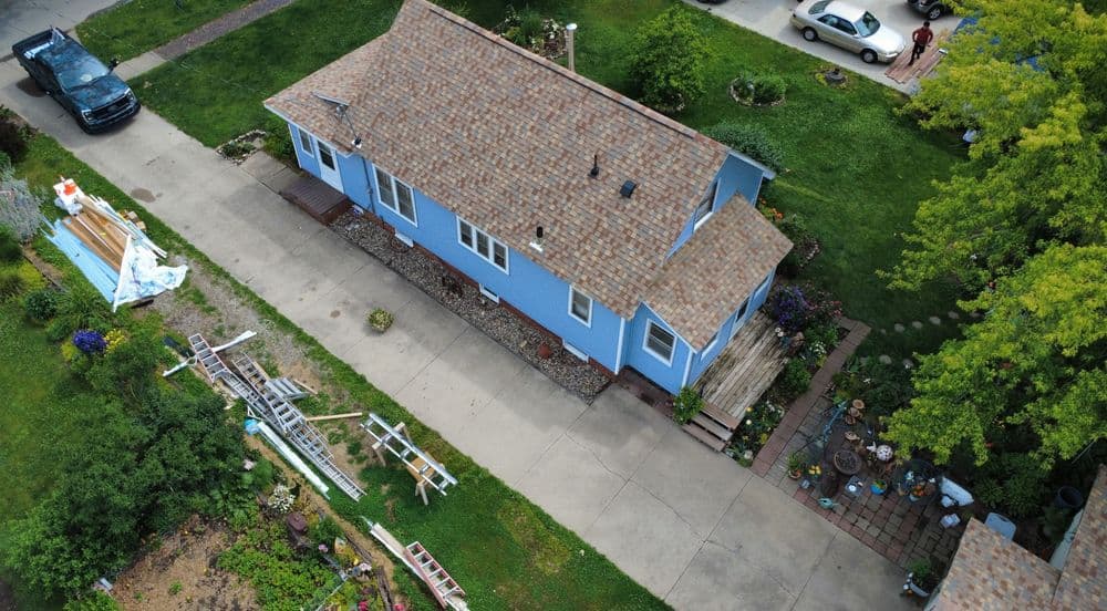 Aerial view of a blue house with a garden and gravel driveway, surrounded by greenery.
