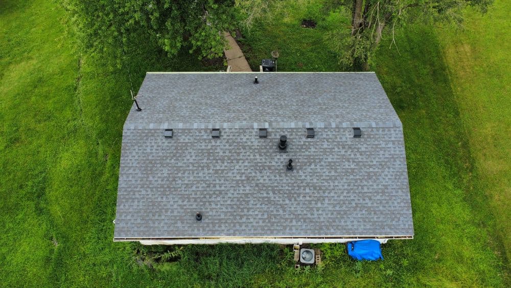 Aerial view of a gray shingled roof with ventilation pipes surrounded by green grass.