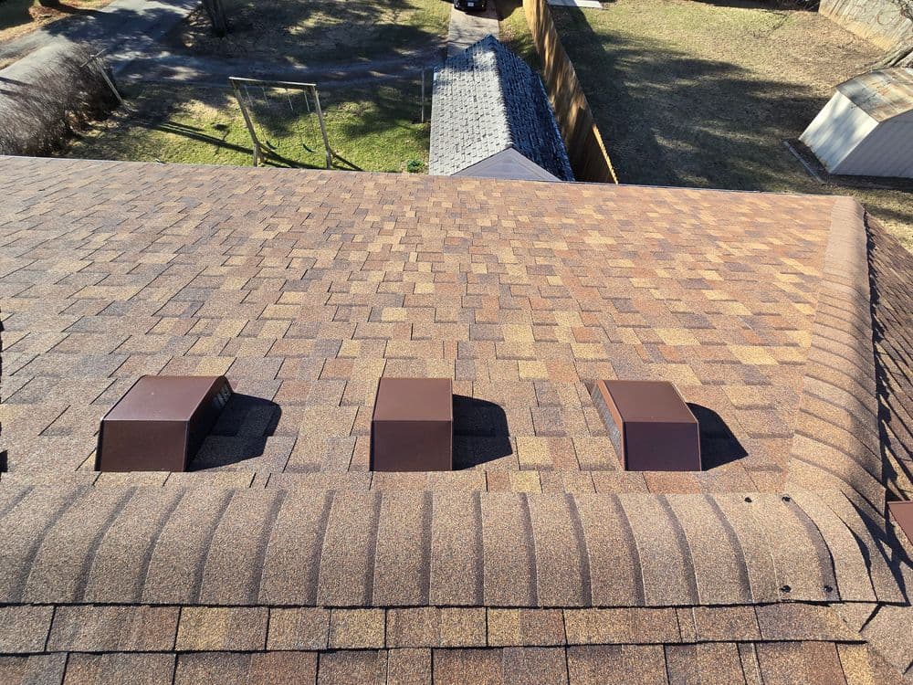 Roof with shingle texture and three brown vent caps, surrounded by green grass and sheds.